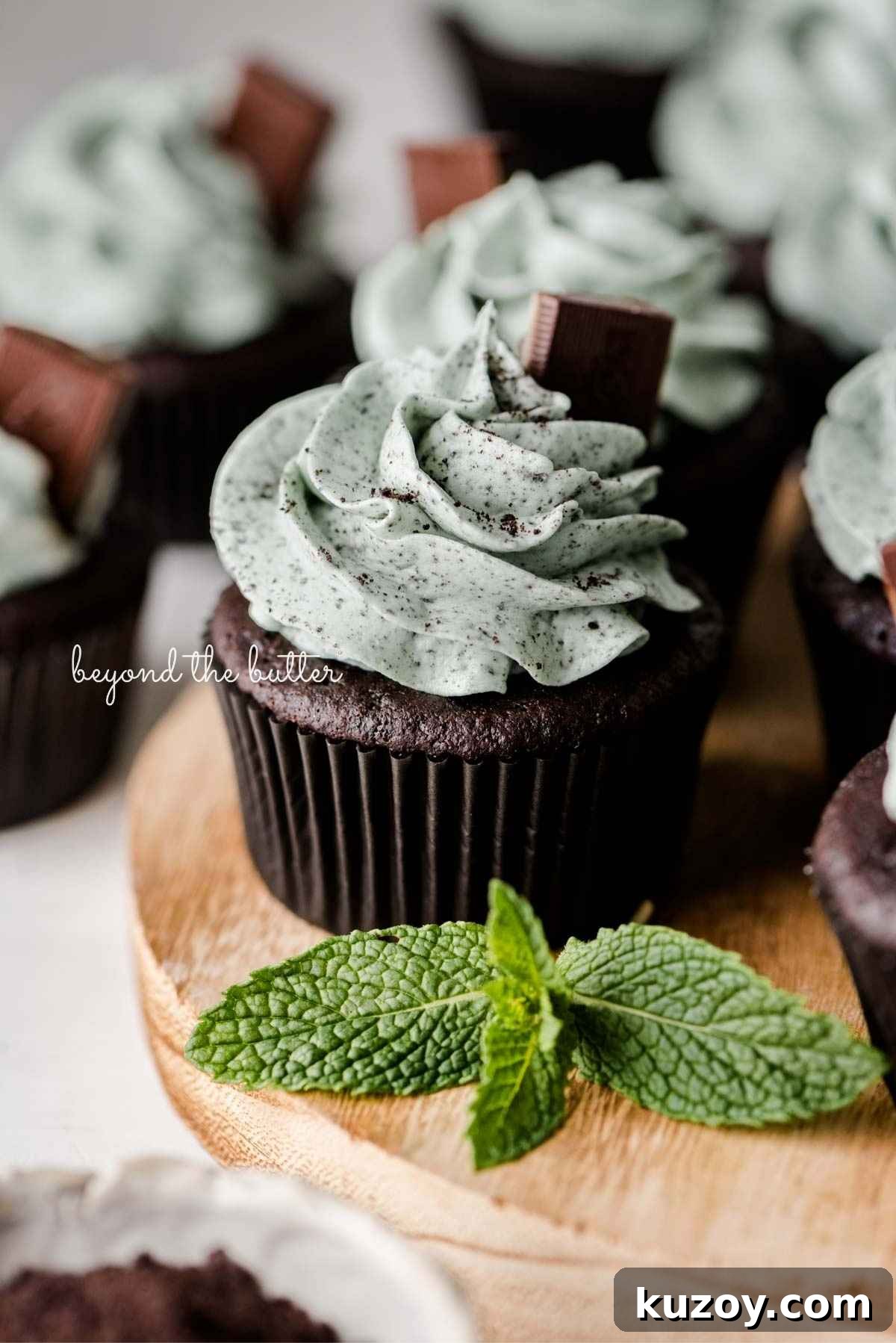 Mint chocolate cupcakes topped with mint chocolate chip frosting and an Andes mint on a wood circular tray with mint leaves as decoration.