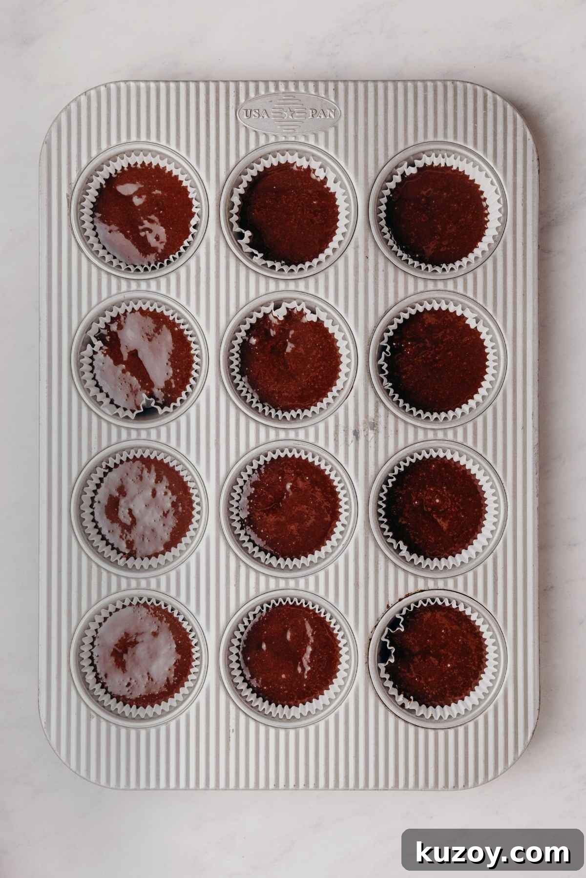 Chocolate cupcake batter evenly divided in metal cupcake tin with cupcake liners on a grey marbled background.