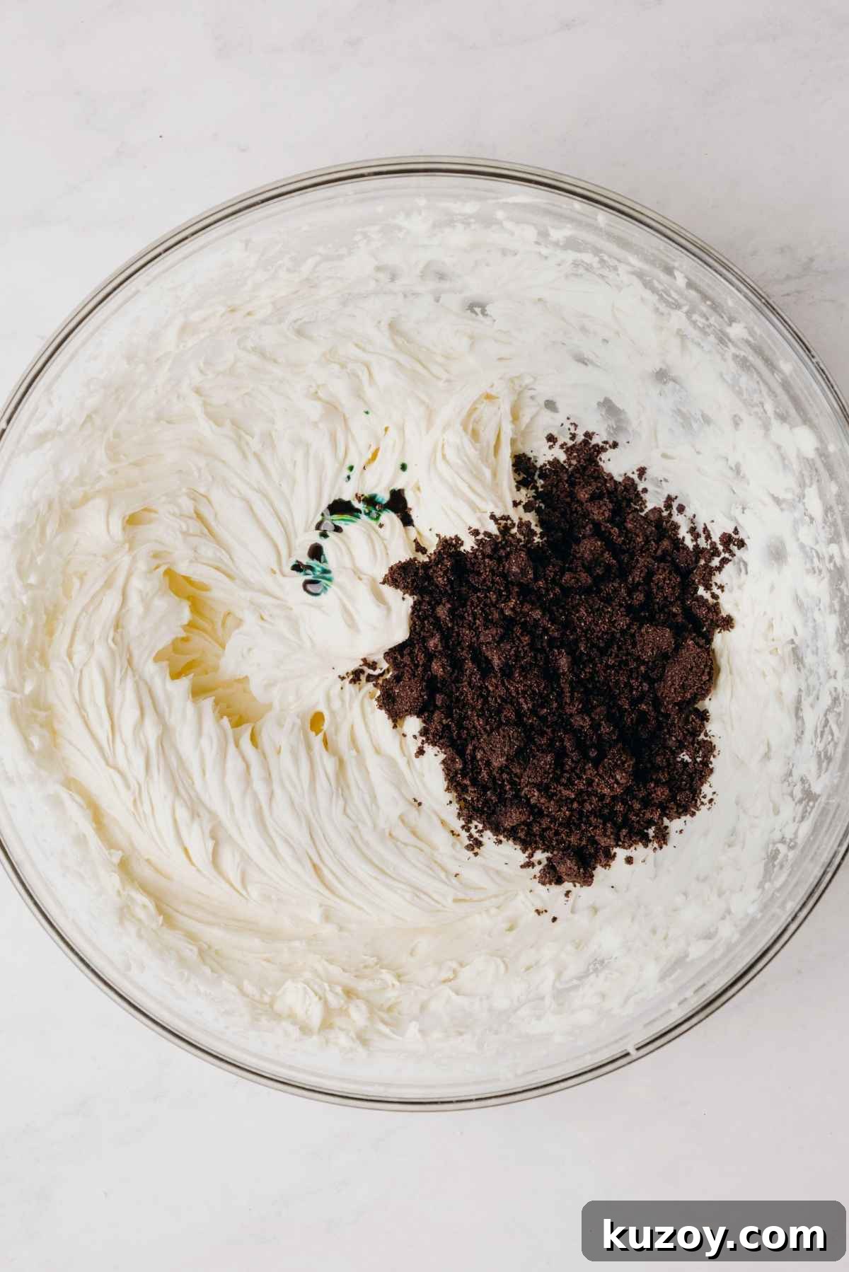 Heavy cream, green food coloring, and crushed chocolate cookies added to the top of mint chocolate oreo buttercream frosting in large glass mixing bowl.