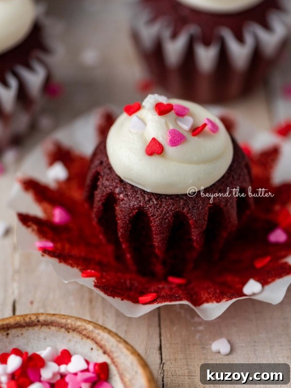 Opened red velvet cupcake with cream cheese frosting topped with heart sprinkles on a light wood background.