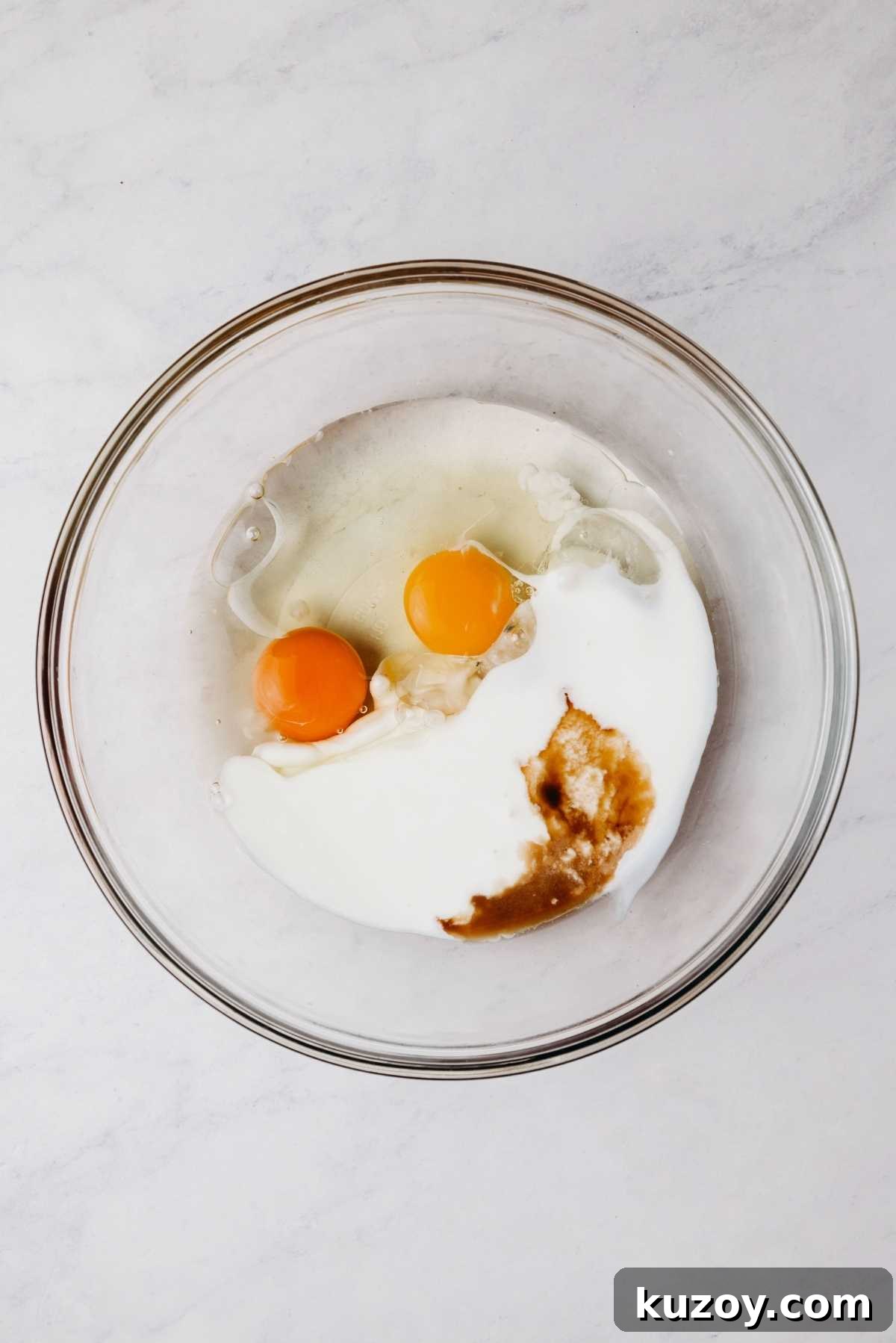 Eggs, buttermilk, vegetable oil, and vanilla extract in a medium size glass mixing bowl on a grey marbled background.