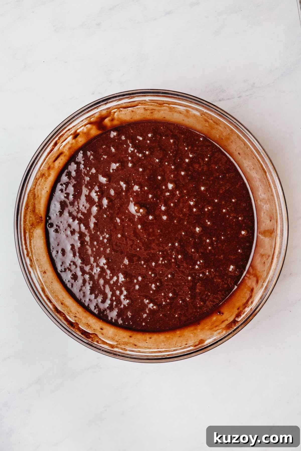 Chocolate cupcake batter fully blended in a medium size glass mixing bowl on a grey marbled background.