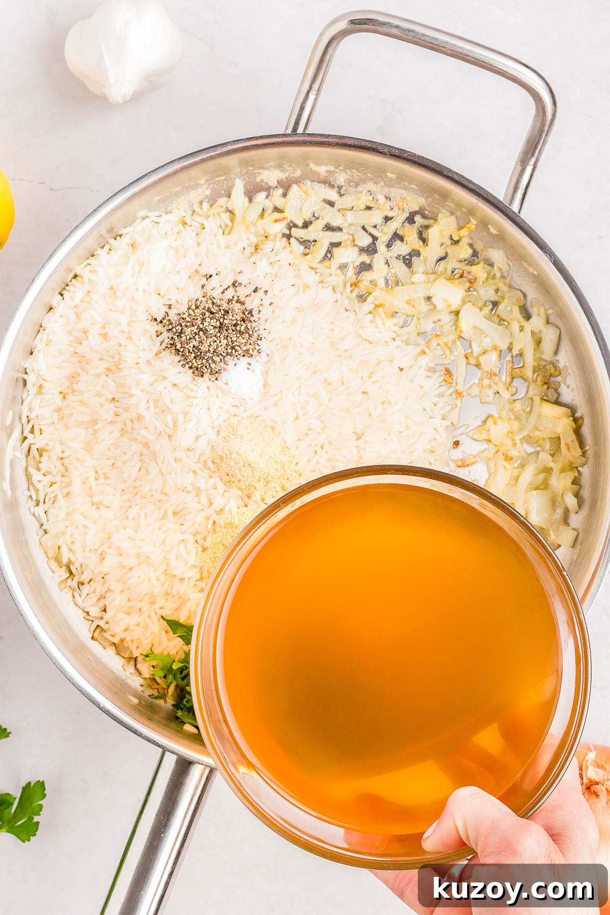 A person pours broth into a pan containing rice, chopped onions, seasoning, and herbs.