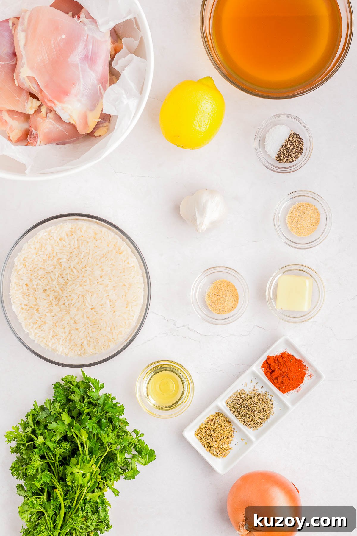 Ingredients for a recipe displayed: chicken, lemon, broth, rice, garlic, onion, spices, herbs, butter, oil, salt, and pepper on a white surface.
