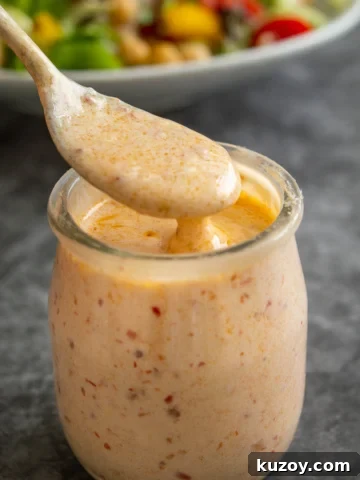 A silver spoon coated in creamy orange dressing over a small glass jar of it on a grey background.