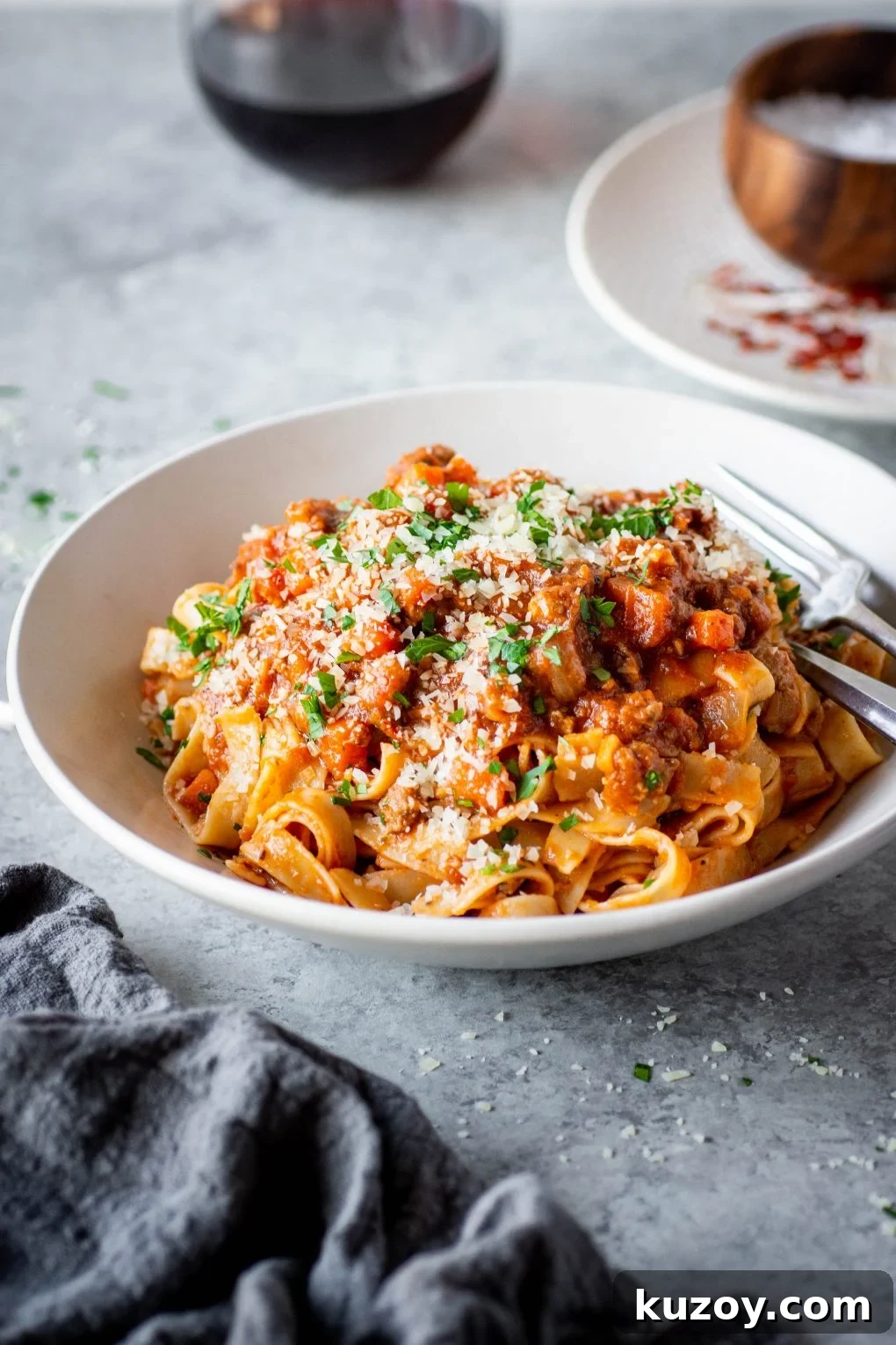 Pumpkin Ragu Pasta Sauce with Canned Pumpkin 2 Side view of a large white bowl filled with fettuccine pasta and pumpkin ragu sauce. Pasta is topped with parmesan cheese and fresh parsley. It is next to a bowl of sea salt and a glass of red wine on a dark grey background.
