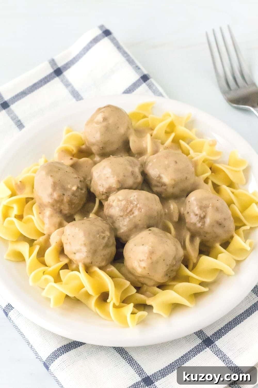 Instant Pot Swedish Meatballs Recipe: Creamy Sauce in 30 Minutes 9 pressure cooker swedish meatballs served over egg noodles on a white plate, resting atop a blue and white napkin with a fork in the background