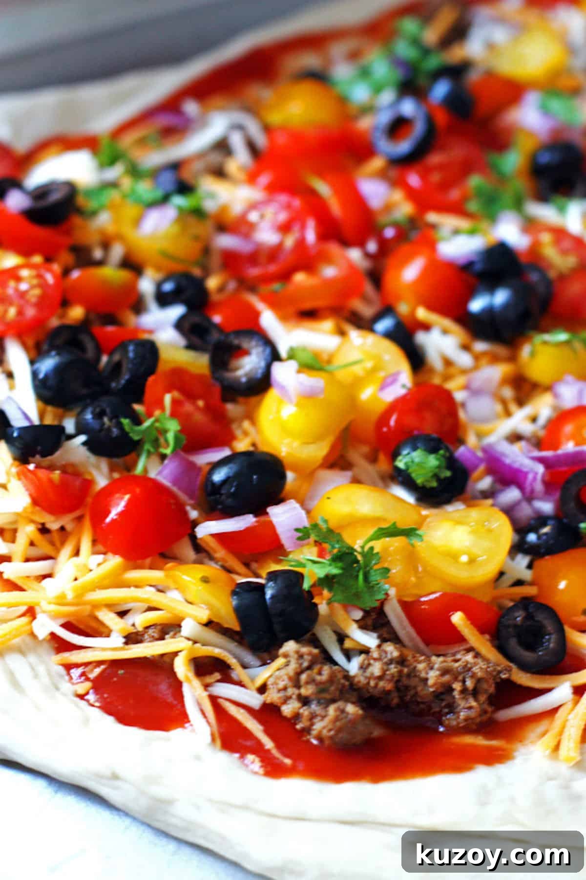 Taco Pizza Recipe: How to Make Homemade Taco Pizza from Scratch 4 An unbaked taco pizza topped with cheese, tomatoes, ground beef, red onions, and black olives.