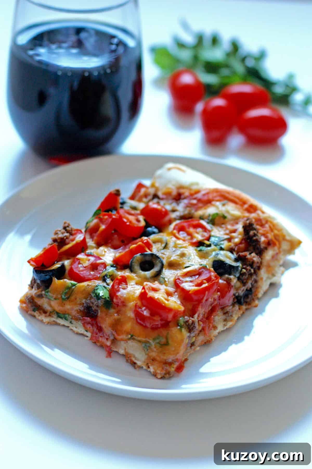 Taco Pizza Recipe: How to Make Homemade Taco Pizza from Scratch 6 A square slice of taco pizza topped with cheese, black olives, and tomatoes, with a glass of wine behind it.