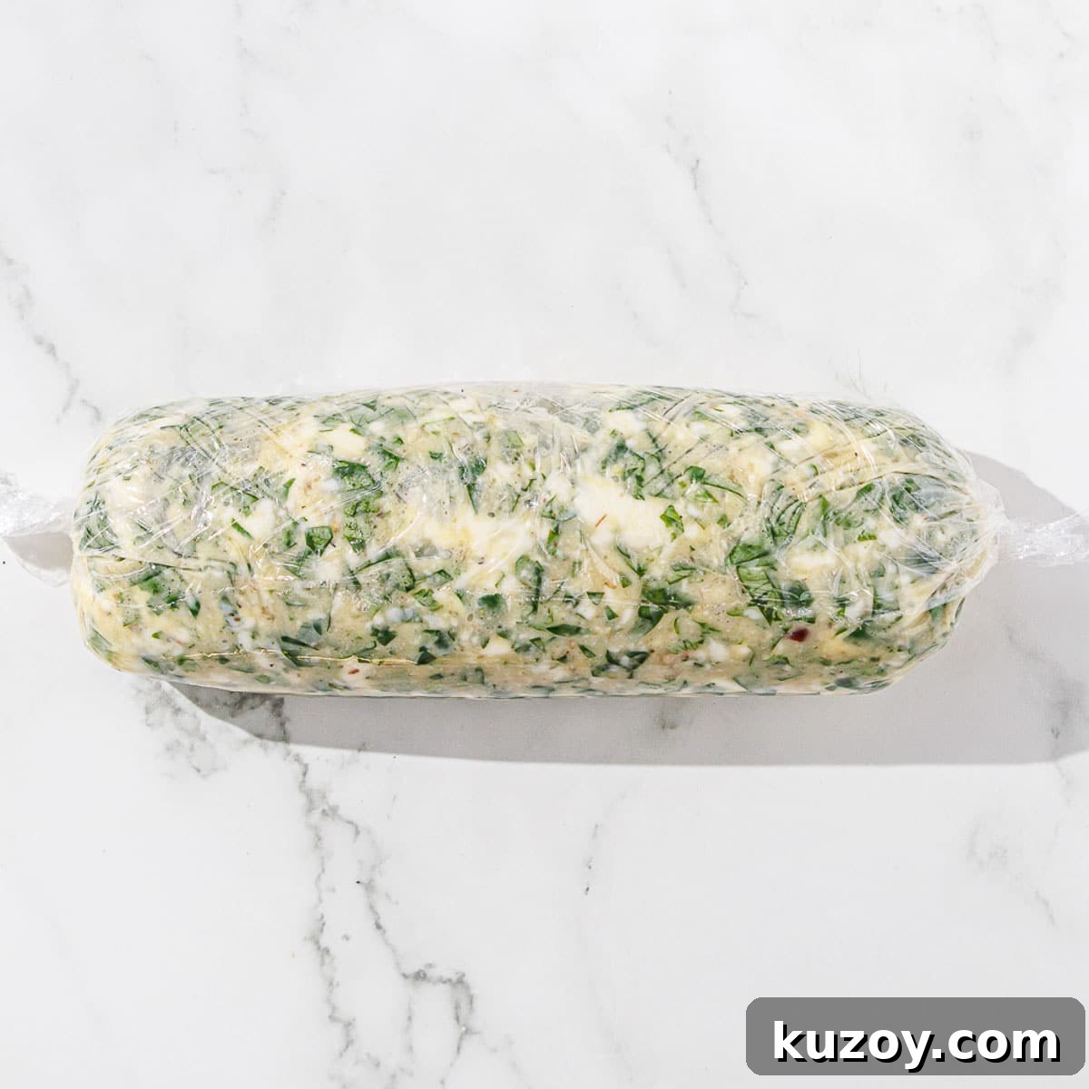 A roll of chimichurri butter wrapped in plastic on a marble surface.