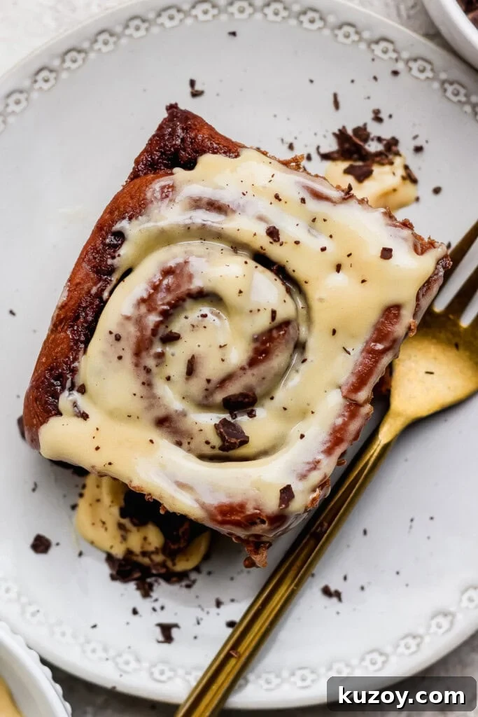 Gooey Chocolate Cinnamon Rolls with Melty Chocolate Center 2 A cinnamon roll topped with cream cheese icing and chocolate shavings on a white plate with a gold fork.