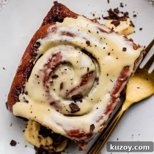 Gooey Chocolate Cinnamon Rolls with Melty Chocolate Center 3 A close-up of a cinnamon roll topped with white icing and chocolate shavings on a plate next to a gold fork.