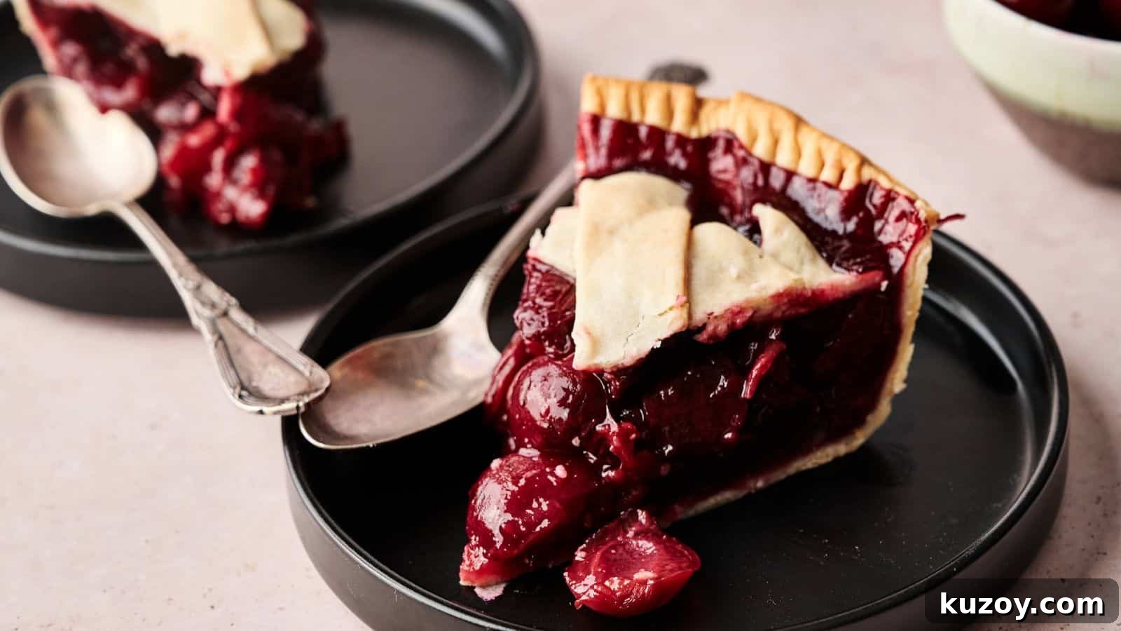33 Must-Bring Thanksgiving Potluck Recipes for Every Year 3 A slice of cherry pie with a flaky crust is served on a black plate with a spoon; another plate with pie is in the background.