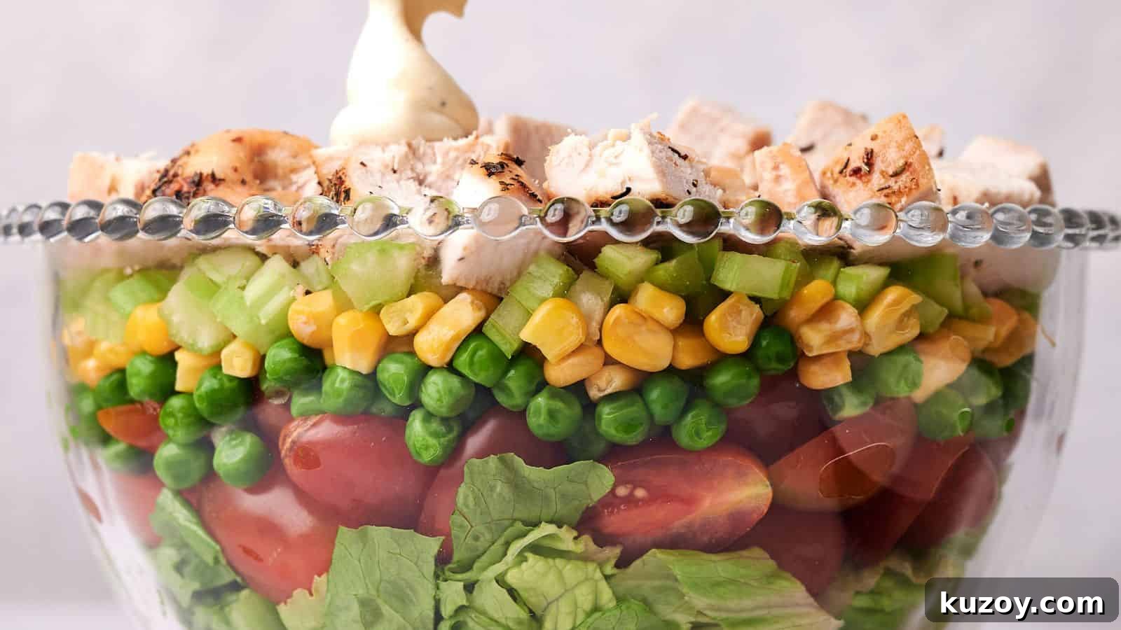 33 Must-Bring Thanksgiving Potluck Recipes for Every Year 31 A clear glass bowl layered with lettuce, cherry tomatoes, green peas, corn, diced celery, and topped with chunks of grilled chicken, garnished with a dollop of mayonnaise.