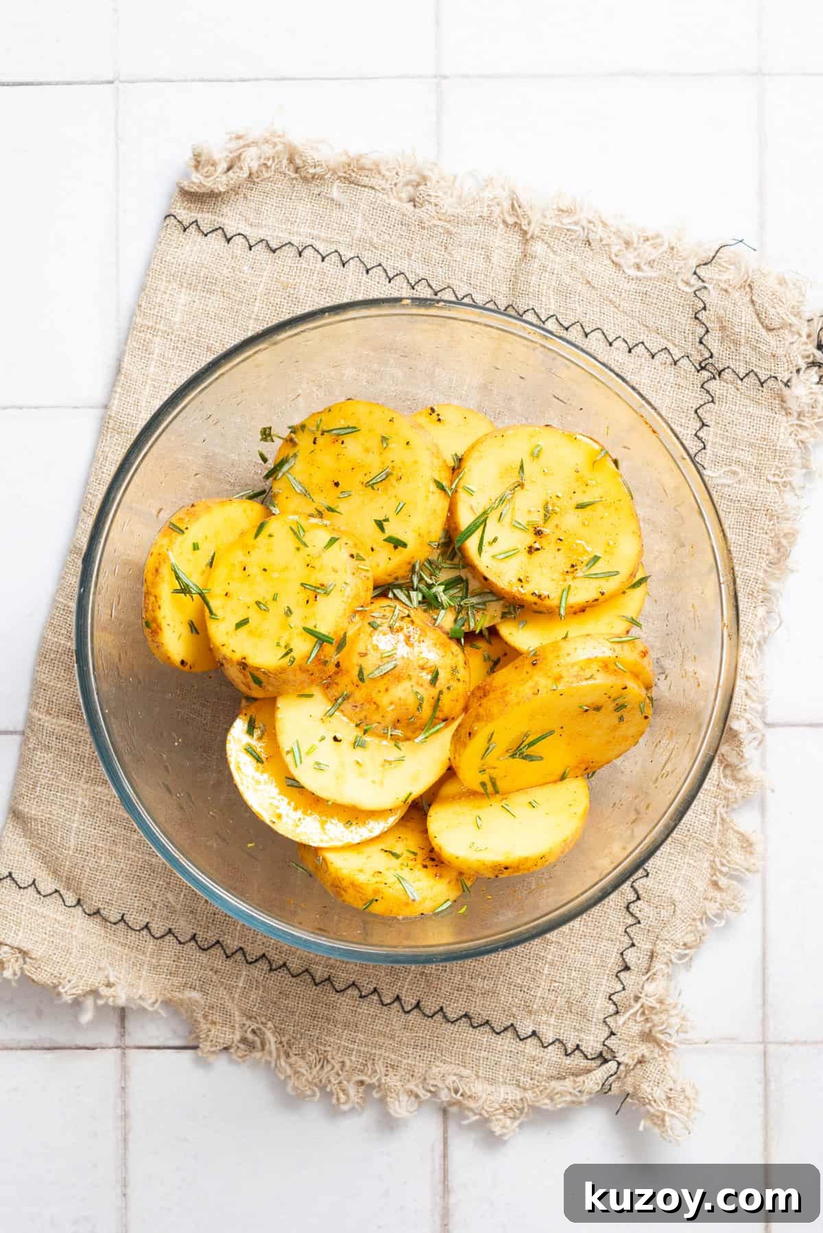 An overhead image of potato rounds in a clear bowl, drizzled with oil and seasonings.