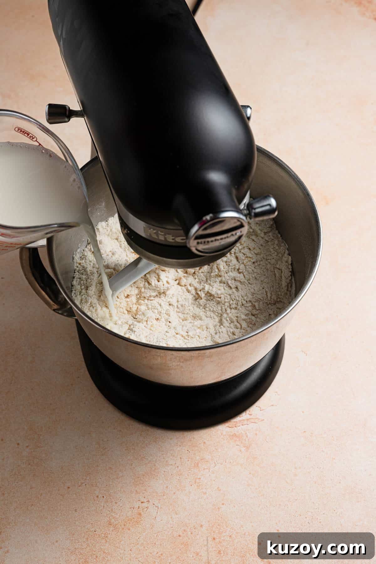 A stand mixer with a bowl filled with flour mixture and milk being poured into the bowl.
