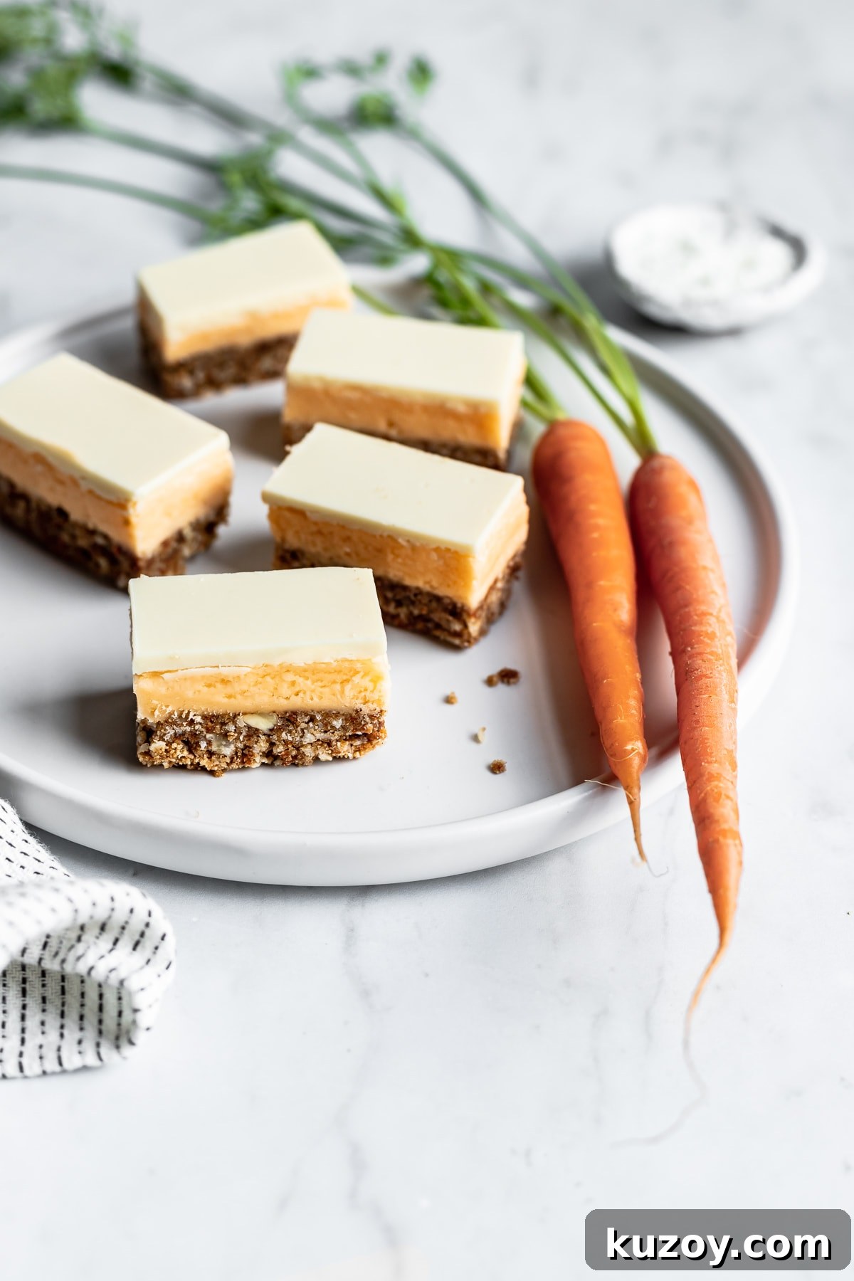 Carrot Cake Nanaimo Bars Recipe — Moist Spiced Dessert Bars 4 Sliced carrot cake bars on a white plate surrounded by carrots, a bowl of coconut on a white marble background