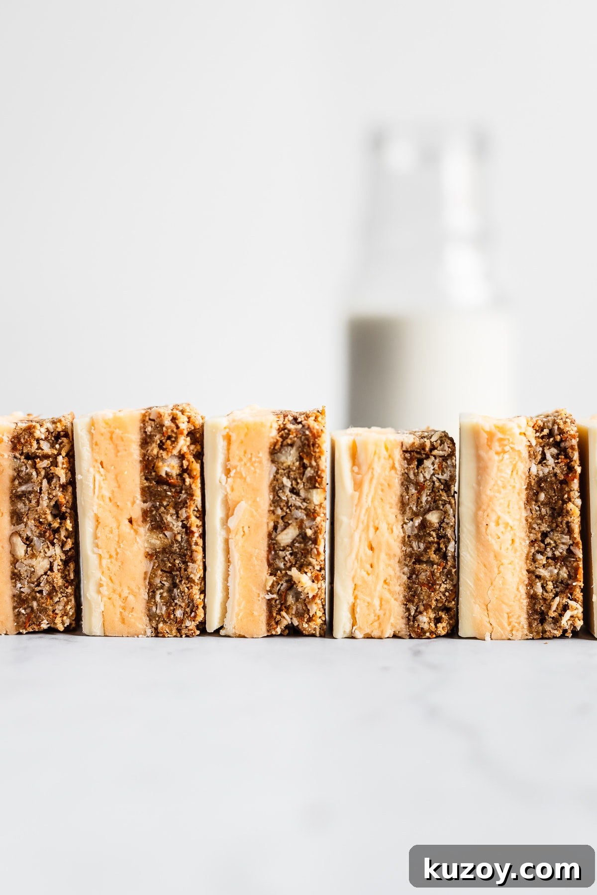 Carrot Cake Nanaimo Bars Recipe — Moist Spiced Dessert Bars 5 A row of sliced carrot cake bars turned on their side and lined up like dominos on a white marble surface with a glass bottle of milk in the background
