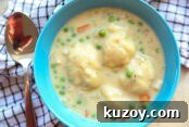 A blue bowl filled with creamy chicken and dumplings. A white and blue striped checked kitchen towel is wrapped around the bowl from the bottom left to the upper right of the bowl. A spoon is on top of the towel on the left side of the bowl.
