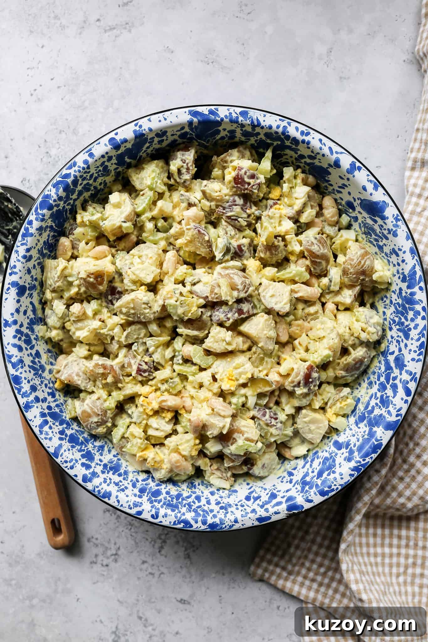 high protein potato salad in a blue enamelware bowl next to a tan gingham kitchen towel and a large serving spoon