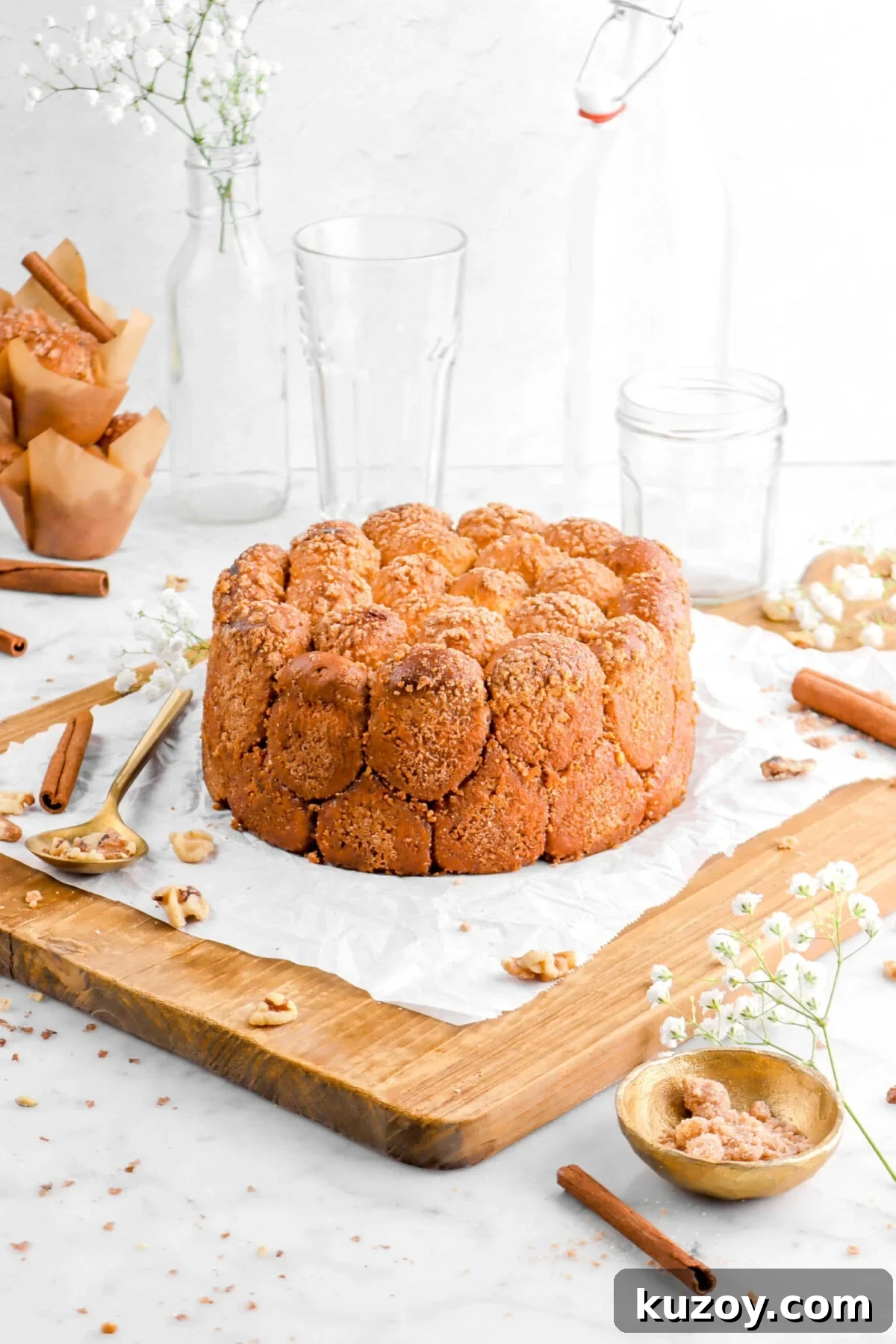 Cinnamon Sugar Walnut Pull-Apart Bread Recipe 2 cinnamon sugar walnut pull apart bread on crumbled parchment paper on a wood board with walnuts, cinnamon sticks, a gold spoon and bowl beside, a stack of muffins, an empty glass, and flowers behind