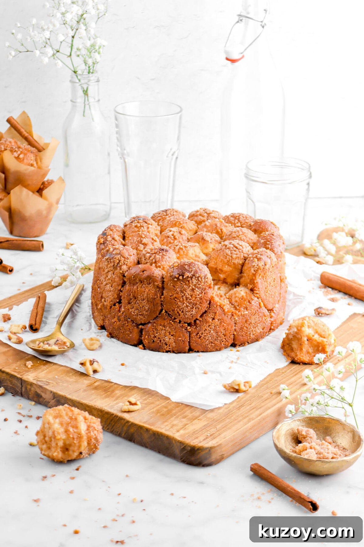 Cinnamon Sugar Walnut Pull-Apart Bread Recipe 25 pulled back shot of cinnamon sugar pull apart bread on wood board with two pieces laying in front, a gold bowl and spoon beside, cinnamon sticks, flowers, and glasses behind on marble surface