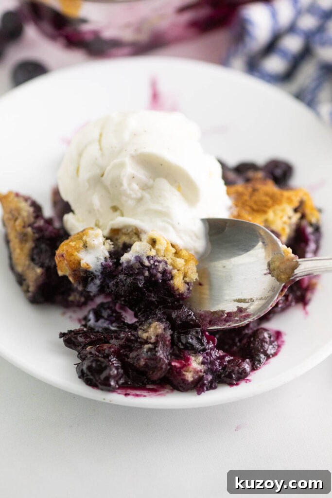 Homemade Blueberry Cobbler Recipe with Golden Crisp Topping 3 Blueberry cobbler on a small white plate with a spoon taking a bite out.