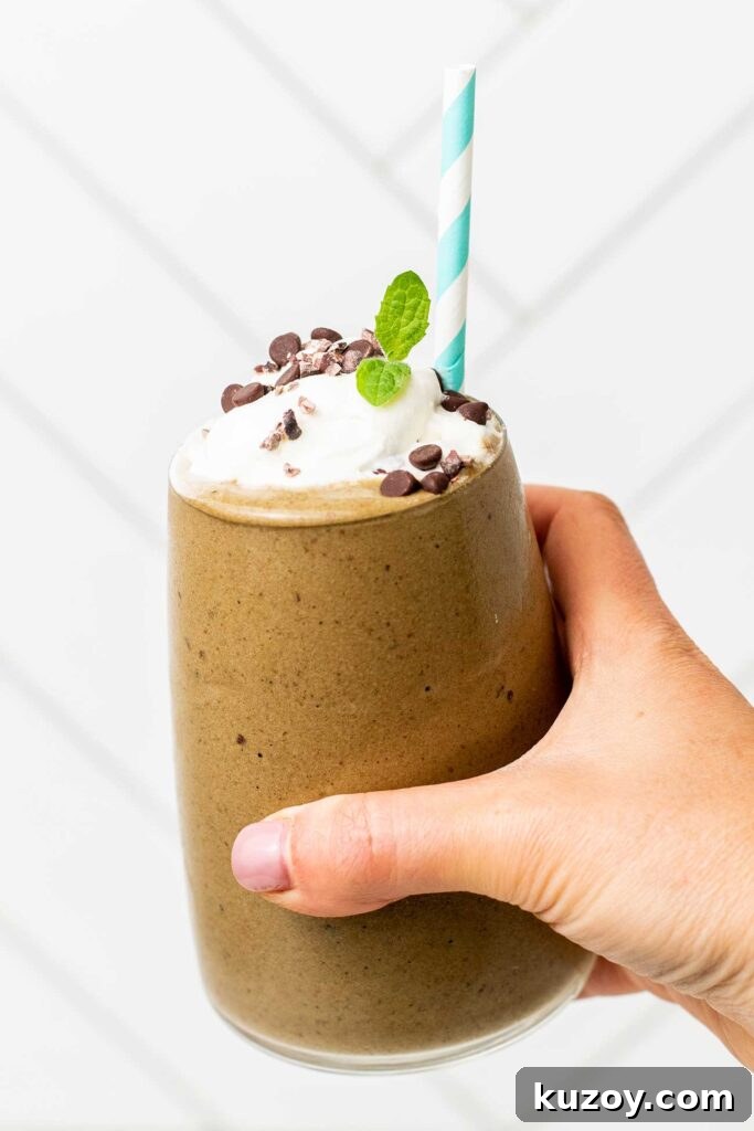 A hand holding a glass filled with a mocha green smoothie topped with whipped cream and cacao nibs.