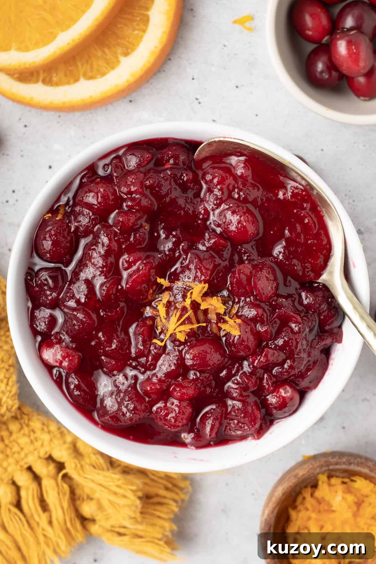 Citrus Cranberry Sauce Recipe: Orange-Infused Holiday Cranberry Sauce 8 A top down shot of a spoon resting in a bowl of cranberry sauce next to a gold cloth and orange slices.
