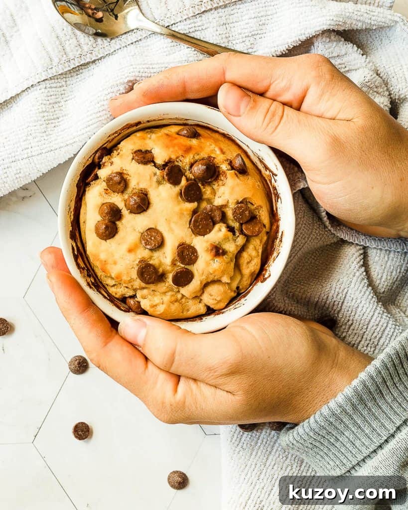 An overhead photo of someone holding a chocolate chip cookie in a mug. There are some chocolate chips and a spoon in the background serving as decoration.