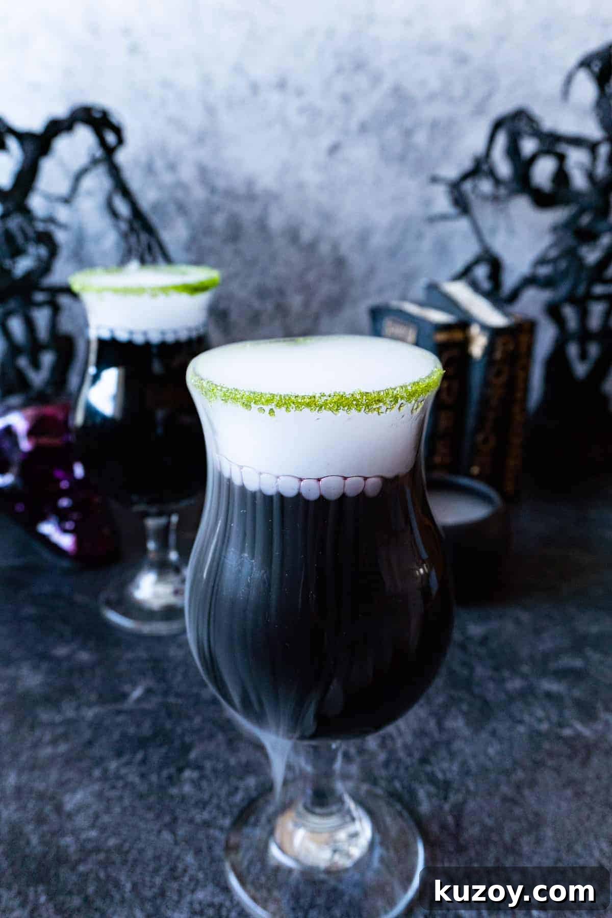 Spooky Cauldron Cocktail Recipe for Halloween Parties 3 black cauldron cocktail in a clear glass cup with a green sugared rim and with dry ice "smoke" spilling out. Halloween decorations in the background.