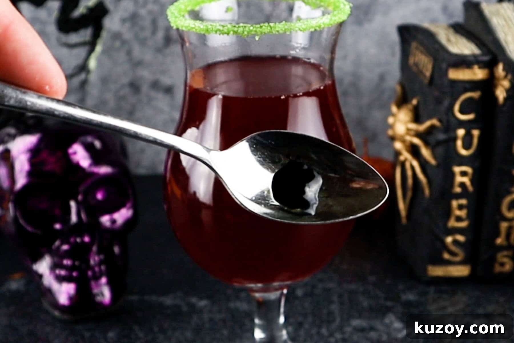 Spooky Cauldron Cocktail Recipe for Halloween Parties 7 pea sized amount of black food gel on a spoon in front of a cocktail being made.