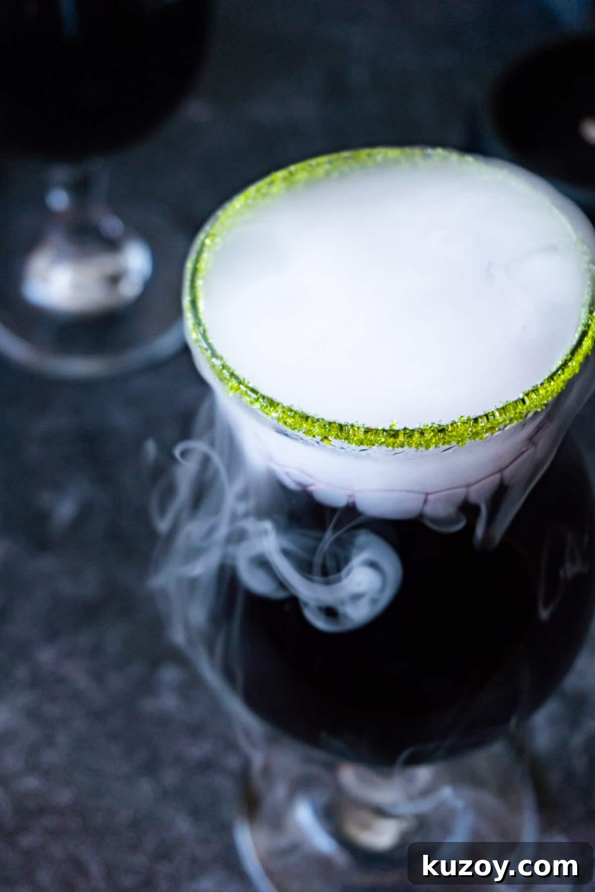 Spooky Cauldron Cocktail Recipe for Halloween Parties 9 View of the top of a black cauldron cocktail in a clear glass cup with a green sugared rim and with dry ice "smoke" spilling out.