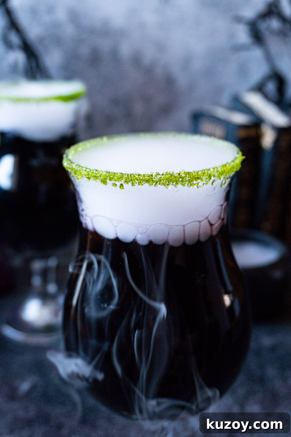 Spooky Cauldron Cocktail Recipe for Halloween Parties 10 black cauldron cocktail in a clear glass cup with a green sugared rim and with dry ice "smoke" spilling out, and Halloween decorations in the background.