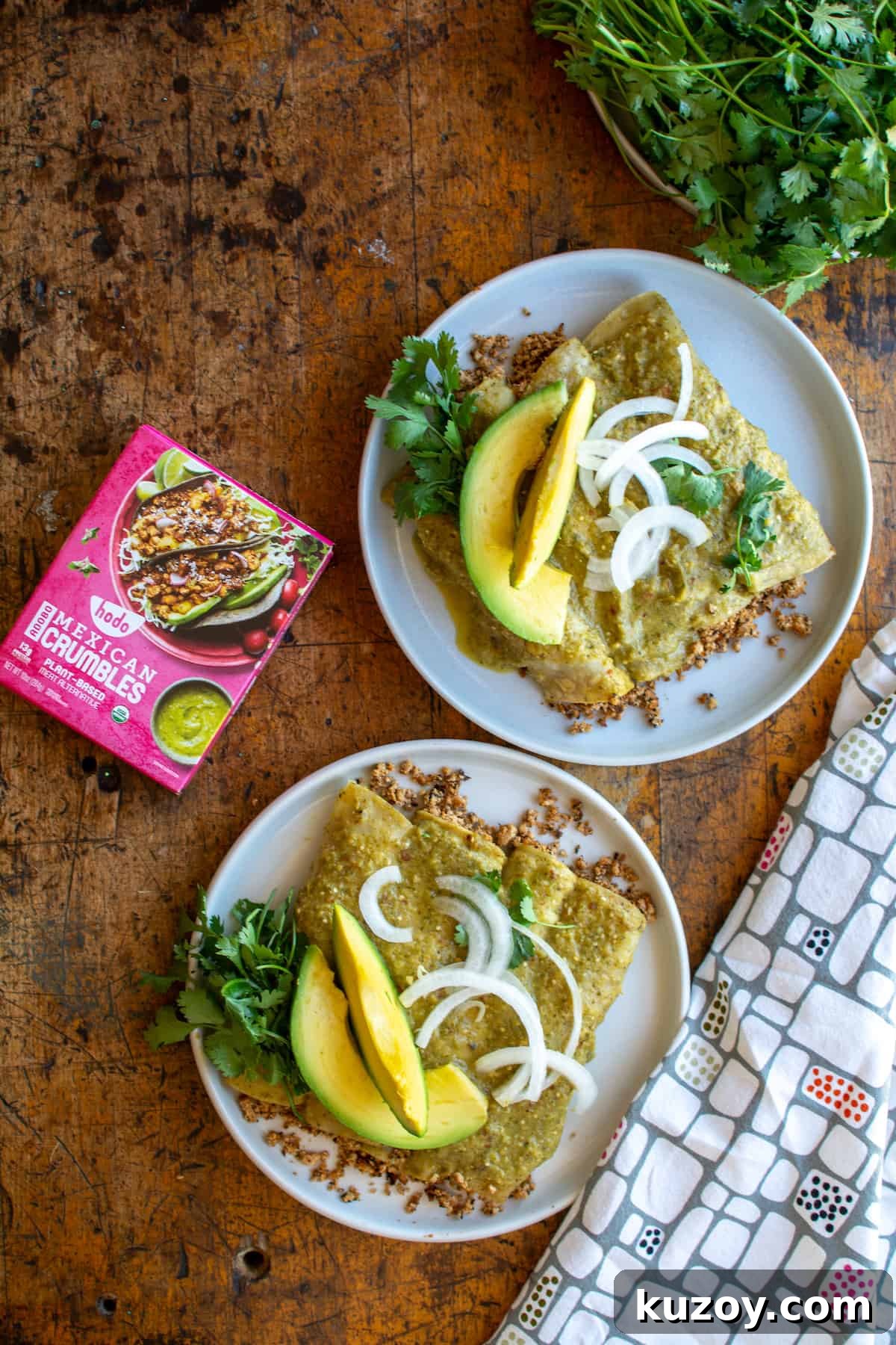 Two plates of enchiladas on a wood table next to a box of Adobo Mexican Crumbles and a bunch of cilantro with a kitchen towel near by.