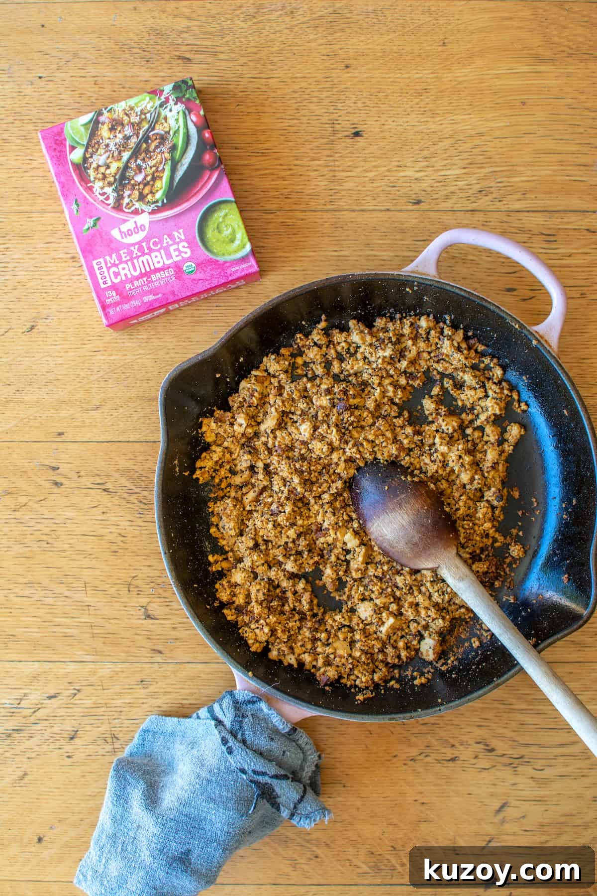 A frying pan filled with browned Hodo Adobo Mexican Crumbles and a wood spoon inside the pan with a box of crumbles next to it.
