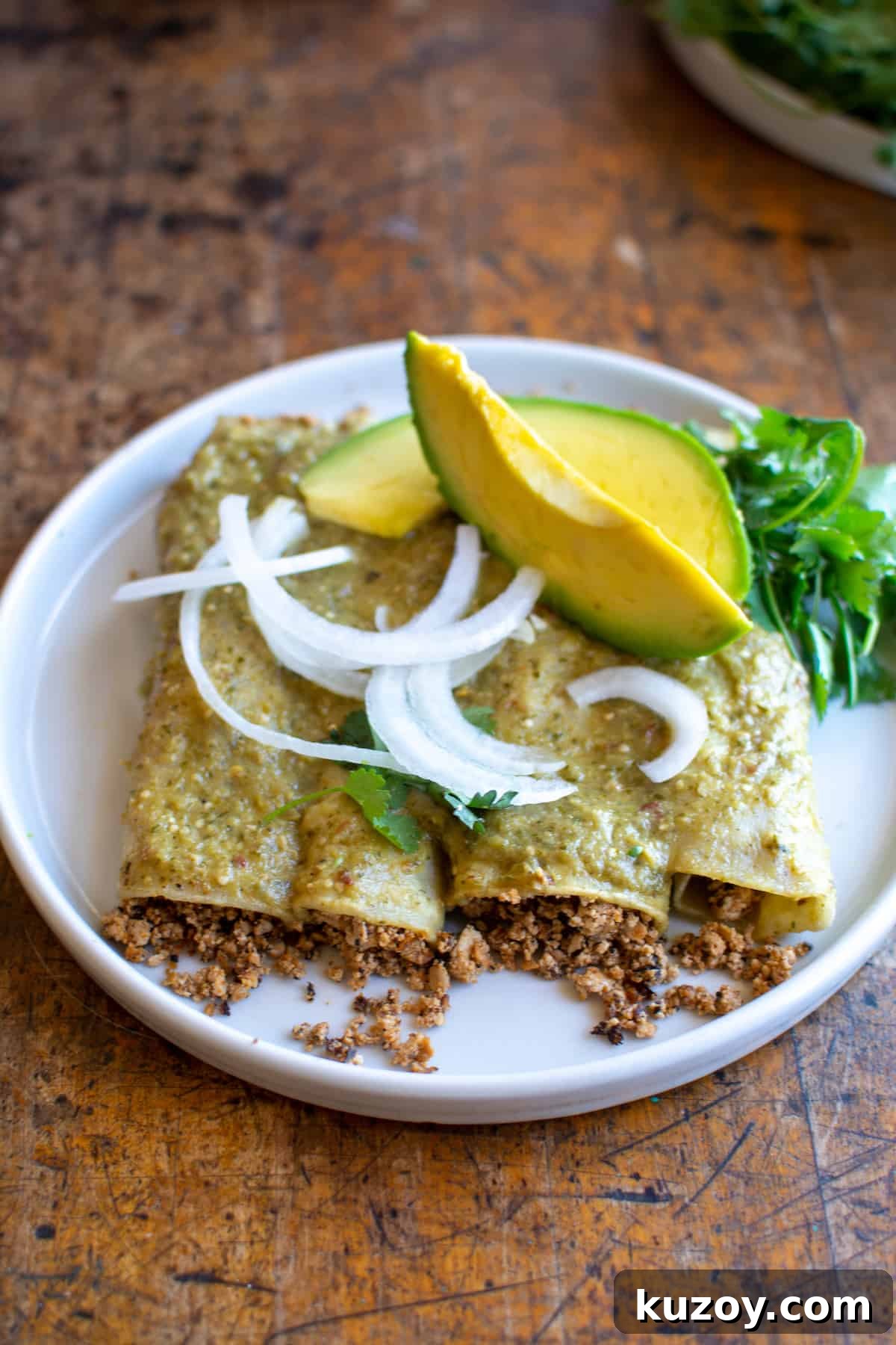 Four vegan enchiladas sitting on a white plate with sliced white onions and avocado on top and a bunch of cilantro on the plate.