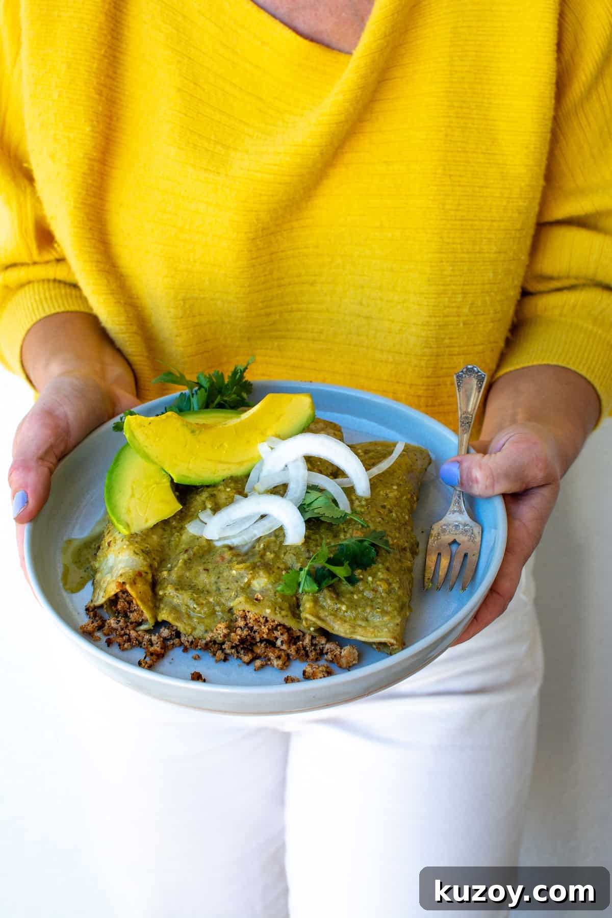 A woman in a yellow sweater and white pants holding a plate of vegan enchiladas and a fork on the side.