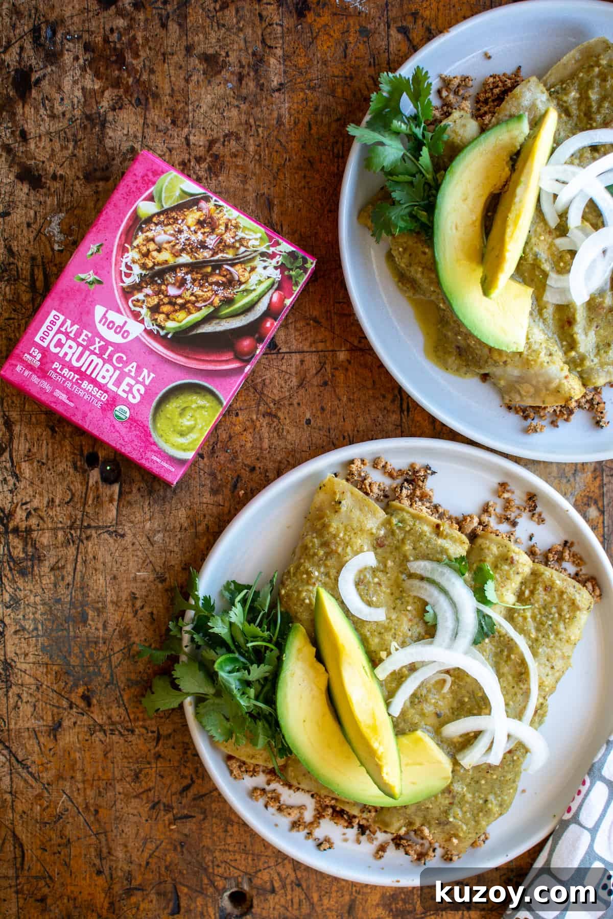 Two plates of enchiladas topped with avocado and onions next to a pink box of Hodo Mexican crumbles