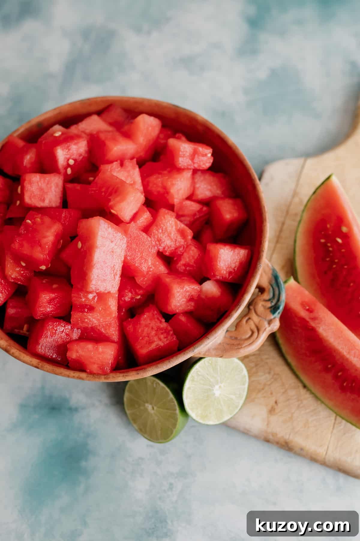 Spicy Jalapeño Watermelon Mezcal Margarita Recipe 4 overhead shot of watermelon cubes in a terracotta bowl with a halved lime and some watermelon wedges on the side.