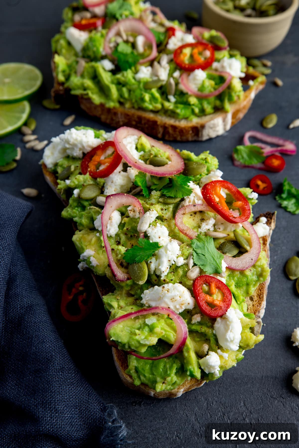 Creamy Avocado Toast with Lemon, Chili Flakes, and Crispy Sourdough 2 Two pieces of loaded avocado toast with feta, shallot, sliced red chilli, seeds and coriander (cilantro) on a dark grey background. There are ingredients scattered around and a dark grey napkin on the left of the frame. There is a pinch pot of seeds at the top right of the frame.
