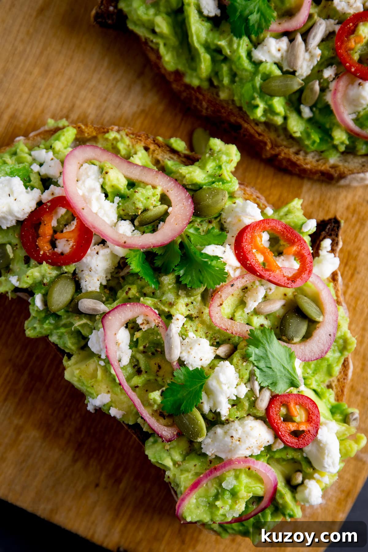 Creamy Avocado Toast with Lemon, Chili Flakes, and Crispy Sourdough 3 A piece of loaded avocado toast with feta, shallot, sliced red chilli, seeds and coriander (cilantro) on a wooden board. There is a further piece, just in shot at the top of the frame.