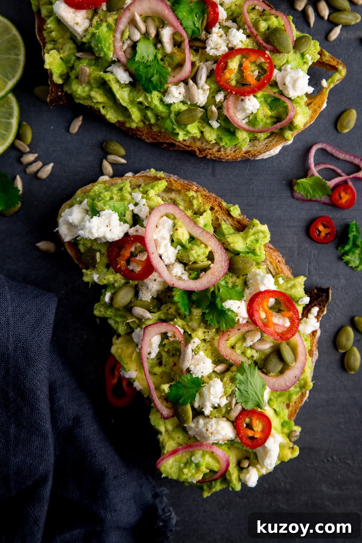 Creamy Avocado Toast with Lemon, Chili Flakes, and Crispy Sourdough 4 Two pieces of loaded avocado toast with feta, shallot, sliced red chilli, seeds and coriander (cilantro) on a dark grey background. There are ingredients scattered around and a dark grey napkin on the left of the frame.