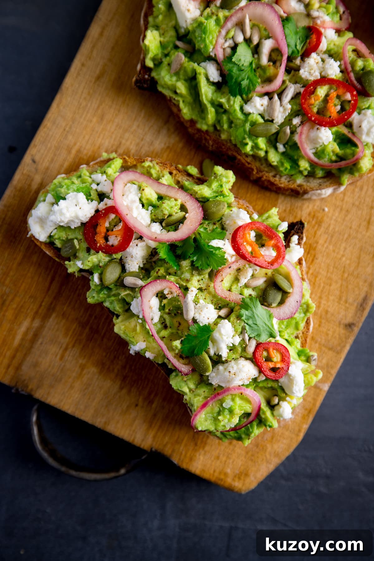 Creamy Avocado Toast with Lemon, Chili Flakes, and Crispy Sourdough 6 Two pieces of loaded avocado toast with feta, shallot, sliced red chilli, seeds and coriander (cilantro) on a wooden board on a dark grey background.