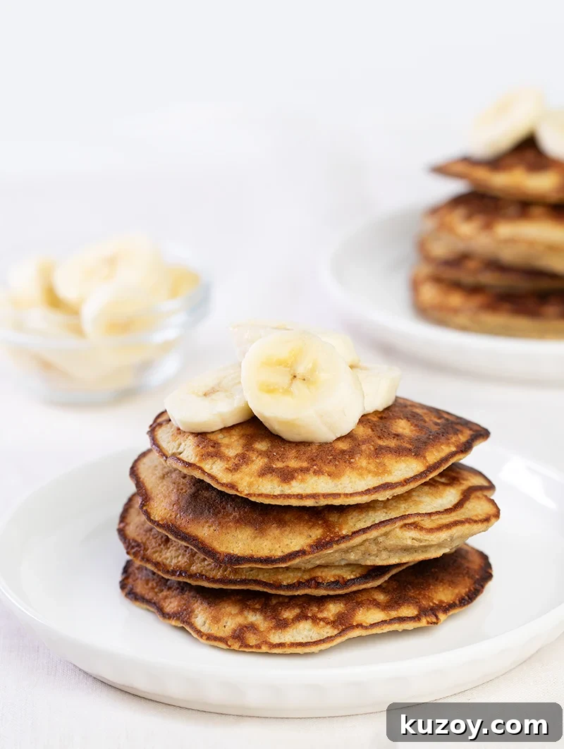 Stack of 4 banana pancakes with banana slices on top on white plate.