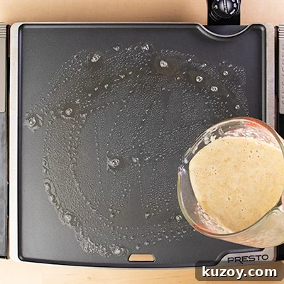 Measuring cup pouring pancake batter onto greased griddle.