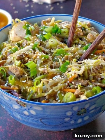 egg roll in a blue flowered bowl with chopsticks and a small white dish of plum sauce in the background and wok with more egg roll in a bowl in the background