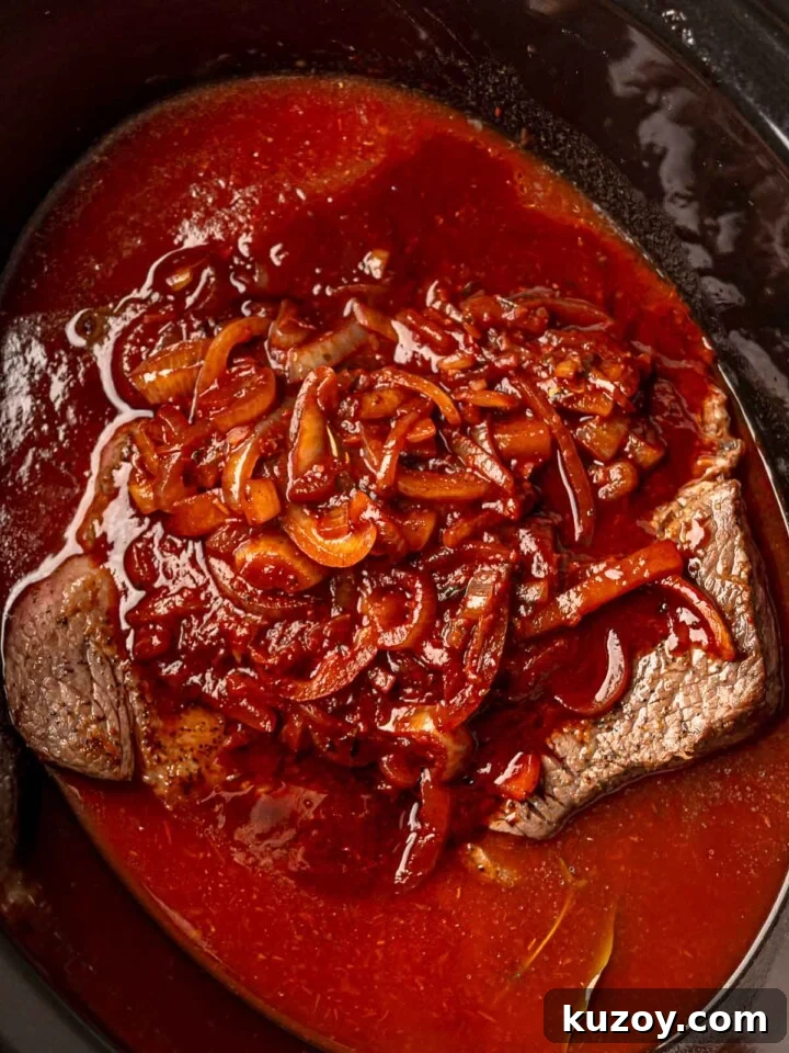 Chuck roast in a slow cooker covered with tomato and red wine sauce and onions.