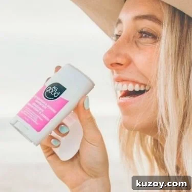 A teenage blonde-haired feminine presenting person wearing a hat is smiling and smelling a natural deodorant stick from All Good.