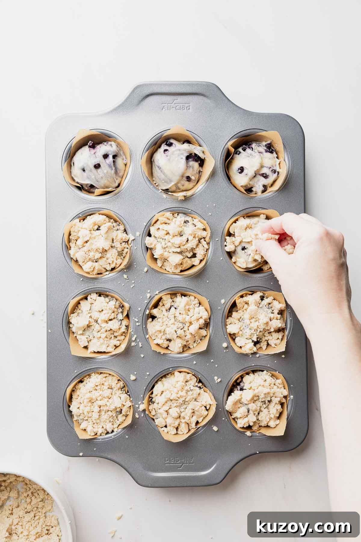 Gluten-Free Blueberry Muffins with Crunchy Streusel Topping 10 Muffin batter divided amongst parchment paper lined muffin tins, hand topping with streusel.